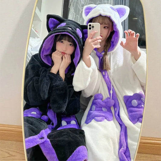 Two women taking a mirror selfie wearing plush cat-themed hooded robes, one in black with purple accents and one in white with purple accents.