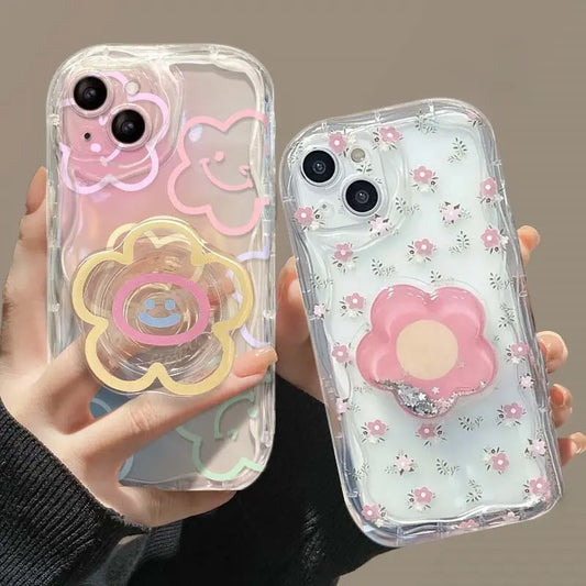 A woman is holding two versions of the Kawaii Cherry Blossom Samsung Phone Case , one design being see through with small floral design and a phone graip in the shape of a cherry blossom flower with floating glitter inside, the other has a more chunky drawn flower design on the case and the phone grip is a chunky drawn flower with a smiley face in the middle.