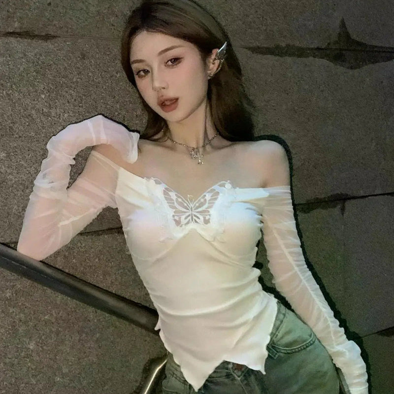 Woman wearing a white lace top and green pants against a gray wall.