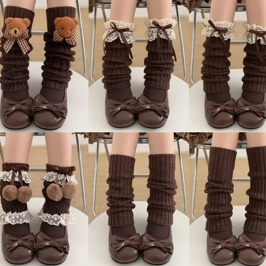 Six pairs of knee-high ribbed leg warmers in brown and gray, shown on a model's legs paired with brown bow flats. The leg warmers feature various decorative details including lace ruffles, pom-poms, hearts, and bows.