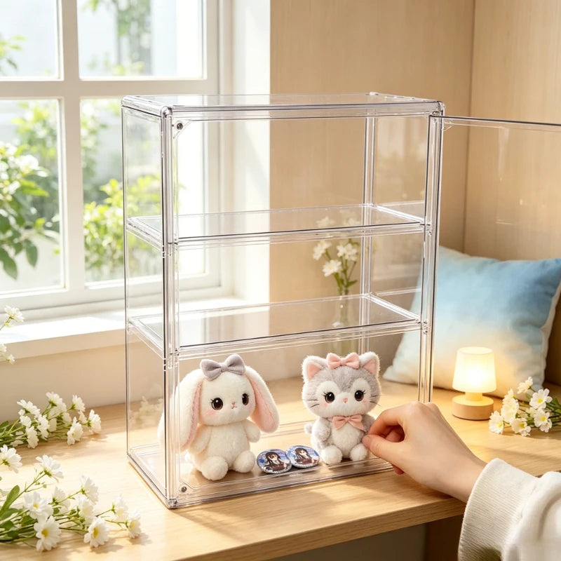A clear acrylic shoe organizer box with three shelves stands on a wooden table in a bright room. Two small plush toys sit inside the organizer, and a person's hand is reaching in to adjust them.