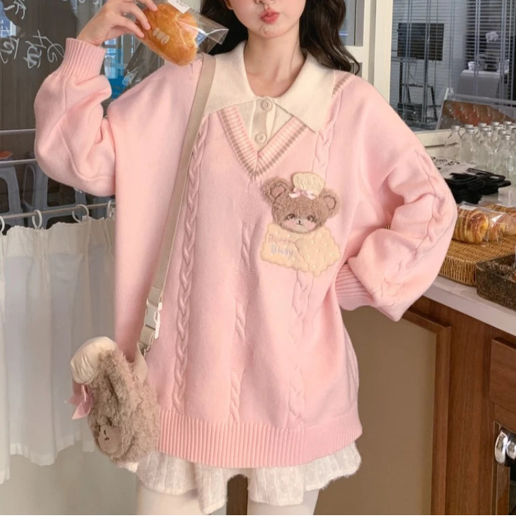 Person wearing a pink sweater with a bear design, holding a pastry in a casual setting.