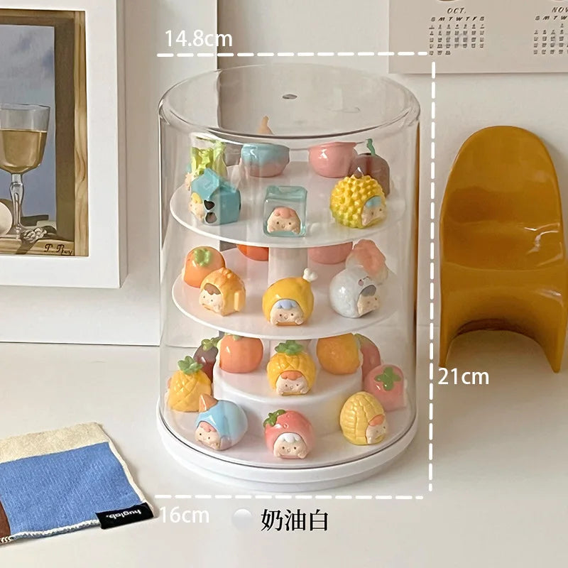 A three-tier rotating acrylic display stand with a clear cylindrical top and white base, shown holding small pastel fruit-shaped figurines. White dashed measurement lines and text labels indicate dimensions: 14.