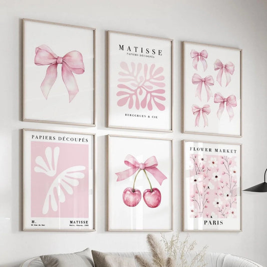 Six framed pastel art prints arranged in a gallery wall above a sofa. The prints feature pink bows, botanical motifs, cherries, and floral text including 'MATISSE' and 'PAPIERS DECOUPEES'.