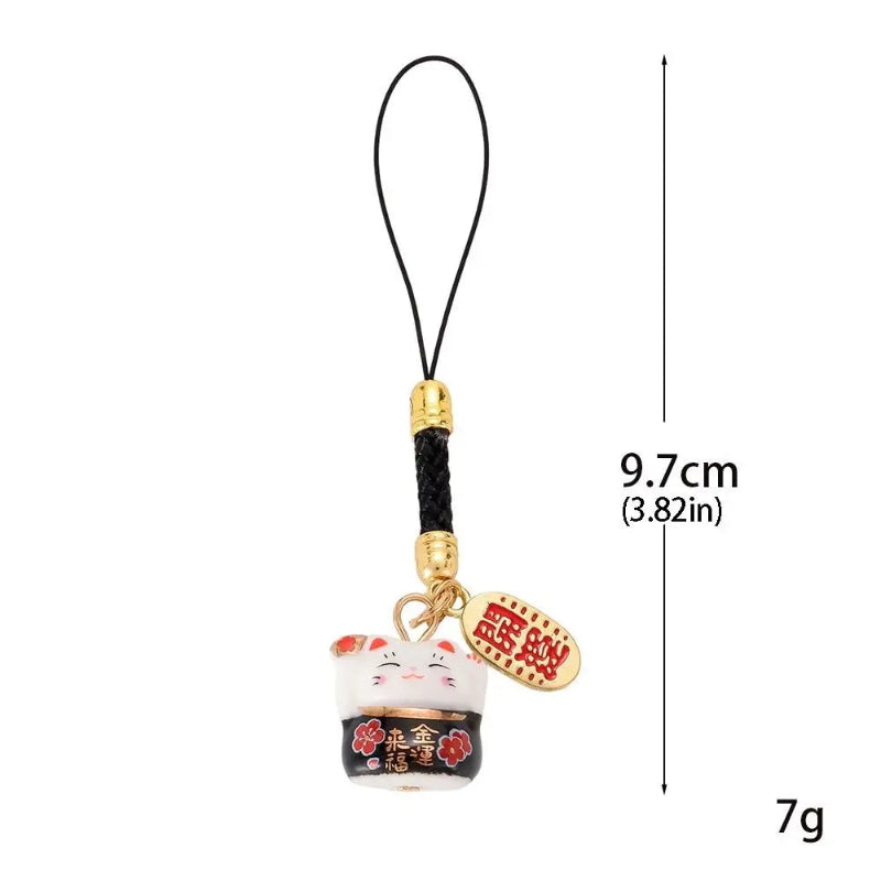 A Japanese maneki-neko lucky cat phone strap charm with a black cord loop, gold-toned metal connector, black braided cord, and a white lucky cat figurine with red and gold details. A dimension marker indicates 9.