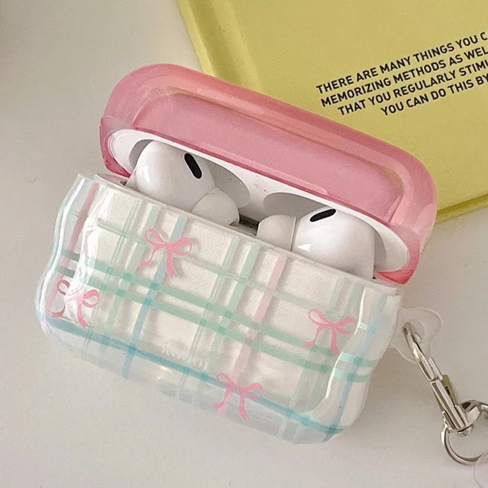 Clear AirPod case with pink lid and plaid design, accompanied by a keychain with a cartoon character, on a light surface.