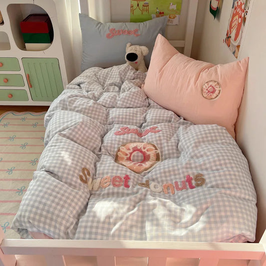 A cozy bed setup featuring a light blue and white gingham checkered comforter with a pink donut graphic and 'Sweet donuts' embroidery, paired with a solid pink pillow and a gray pillow.