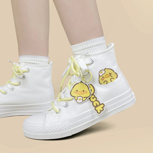 White high-top canvas sneakers with pale yellow laces and cartoon duck charms, shown worn with white crew socks on a plain beige background.