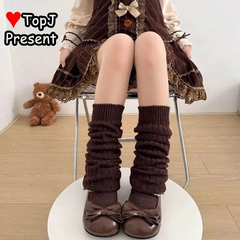 A person seated on a chair wearing dark brown ribbed knee-high socks with matching bow shoes, shown from the waist down in a styled indoor setting. A teddy bear sits on the floor in the background, and the text 'TopJ Present' appears in the upper left.