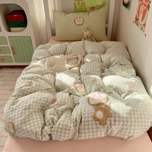 A cozy bed setup in a child's bedroom featuring a light green and white gingham quilted comforter with teddy bear appliques, a matching pillow, and a small plush bear toy.