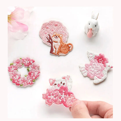 Kawaii Cherry Blossom Iron-On Embroidery Patches come in  four different designs , one with a shiba inu sat next to a cherry blossom tree, another with a white bird flying with a cherry blossom flowers positioned on its wings, also a cat waving and a row of cherry blossom flowers in front , and the last one is the cherry blossoms in a wreath formation. all on a white background.