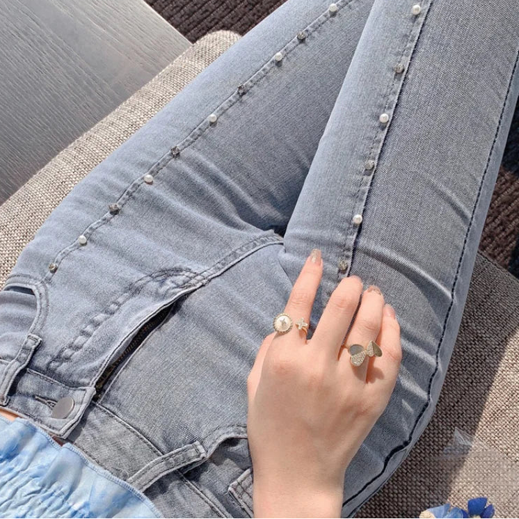 Close-up of a person wearing Voostao light blue denim jeans with pearl embellishments along the seams, seated on a textured couch. A hand wearing silver rings rests on the thigh.
