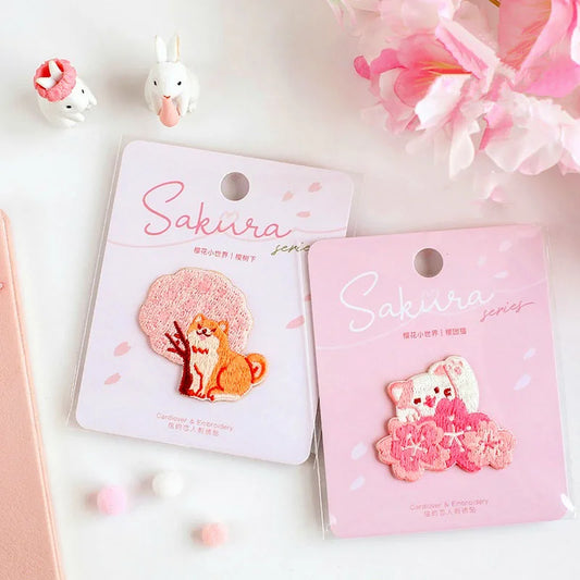 Kawaii Cherry Blossom Iron-On Embroidery Patches come in  four different designs , one with a shiba inu sat next to a cherry blossom tree,  also a cat waving and a row of cherry blossom flowers in front , this is displayed on a white and pink background. with them individually packed in kawaii packaging.