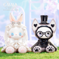Two kawaiii characters, one resembling a rabbit and the other a dog with glasses, standing on grass with a blurred background.