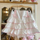 White skirt with pink trim hanging on a wooden hanger against a neutral background.