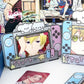 Anime-themed Nintendo Switch-style decorative accessories displayed on a tabletop, including a gray controller-shaped holder with a blonde character face, a pink controller-shaped holder with a green-eyed character face, a small character card, and a partial blue controller-shaped item at the bottom right. Background shows illustrated anime characters.