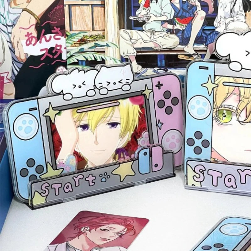 Anime-themed Nintendo Switch-style decorative accessories displayed on a tabletop, including a gray controller-shaped holder with a blonde character face, a pink controller-shaped holder with a green-eyed character face, a small character card, and a partial blue controller-shaped item at the bottom right. Background shows illustrated anime characters.