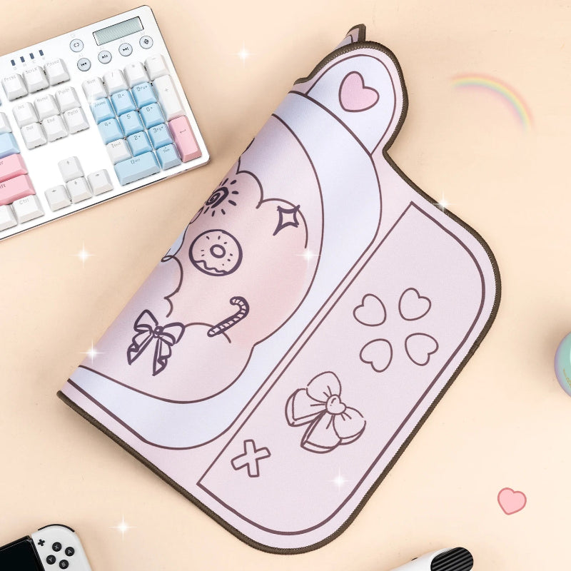 A pastel pink gaming mouse pad with a cute cartoon character design, styled on a peach desk with a keyboard, mouse, and small rainbow sticker.