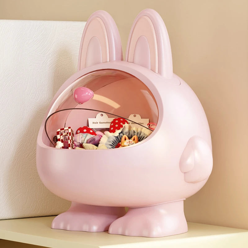 A pink bunny-shaped decorative container with upright ears, a clear dome lid, and small feet at the bottom, shown on a shelf against a beige wall. The open top contains assorted small items.