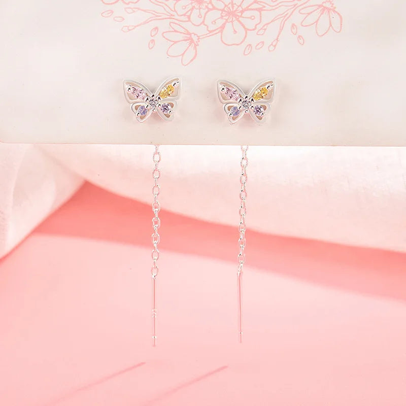 A pair of silver butterfly drop earrings with iridescent pastel gemstones on the wings and long dangling chains, displayed on a soft pink and white background.