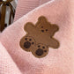 Sweet Teddy Bear Cardigan pastel pink edition with a bear sown into the chest area whilst the sleeves have large black stitches where they have been joined and black strips on the wrist cuffs and black and pink trim, this image is displaying a close up of the bear which is sitting with its arms open.