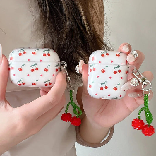 White airpods cases with red cherry pattern held by hands, each with a green beaded keychain.