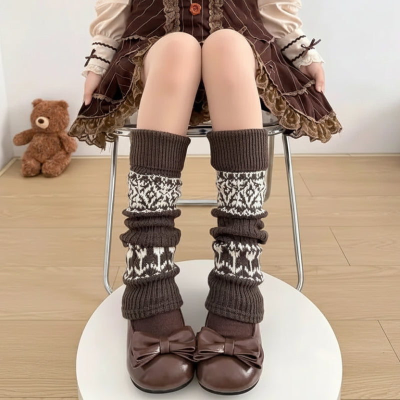 A person wearing long brown ribbed leg warmers with cream and brown geometric knit patterns, paired with brown bow shoes, seated on a metal chair. A 'TopJ Present' logo is visible in the top-left corner.
