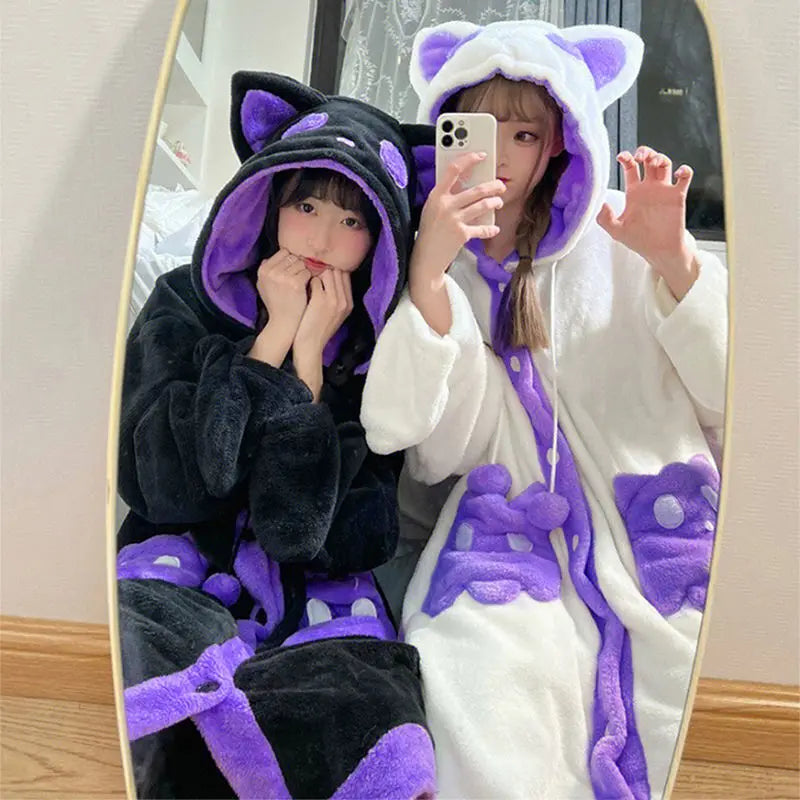 Two women taking a mirror selfie wearing plush cat-themed hooded robes, one in black with purple accents and one in white with purple accents.
