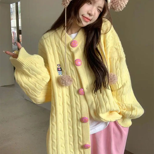 Person wearing a yellow textured cardigan with pink buttons and a matching hat indoors.