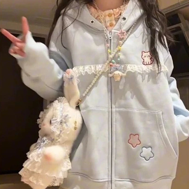 A person wearing a light blue zip-up hoodie with lace trim, pastel embroidered star patches, and a small crossbody bag with a plush bunny and pearl chain, posed indoors with a blurred face.