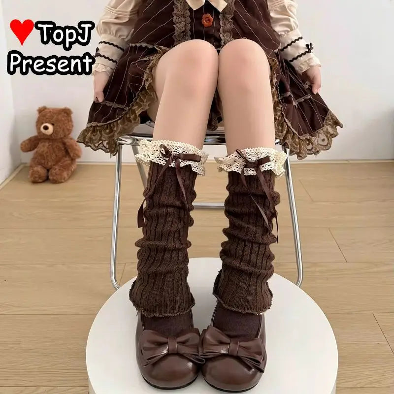 A person seated on a chair wearing long brown ribbed leg warmers with cream lace ruffles and bows, paired with matching brown bow shoes. A teddy bear sits on the floor in the background, and a TopJ Present logo appears in the top left.