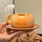 A hand holds a small orange cat-shaped ceramic cup with a matching saucer. The cup has a rounded body, pointed ears, a curved handle, and simple line drawings of a smiling face on the front.