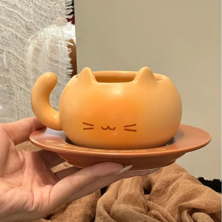 A hand holds a small orange cat-shaped ceramic cup with a matching saucer. The cup has a rounded body, pointed ears, a curved handle, and simple line drawings of a smiling face on the front.