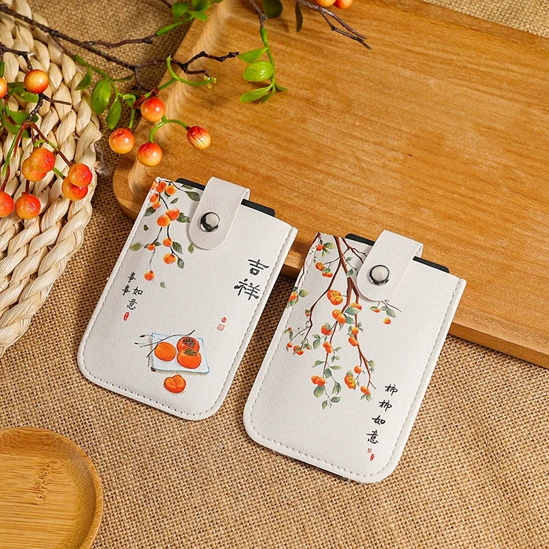 Two white faux-leather phone pouches with snap closures, decorated with orange persimmon illustrations, green leaves, and Chinese calligraphy, displayed on a burlap mat next to a wooden board.