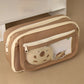 Brown pencil case with a transparent pocket containing a plush toy on a beige surface.