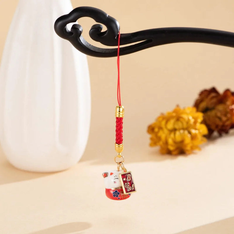A red braided cord charm with a gold-tone connector and a small hanging figurine, displayed on a black decorative stand against a warm beige background with a white vase and yellow flowers.