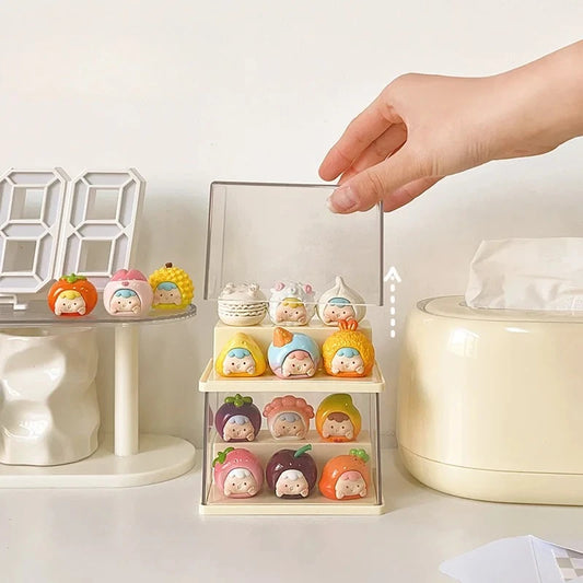 A hand is shown lifting the clear acrylic cover of a small three-tier display stand holding colorful mini character figurines. The stand sits on a white tabletop next to a cream tissue box, a white digital clock, and other small decorative items.
