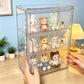 A clear acrylic three-tier display cabinet holding small plush bunny and bear toys, shown on a tabletop with hands holding the cabinet and a softly lit room in the background.