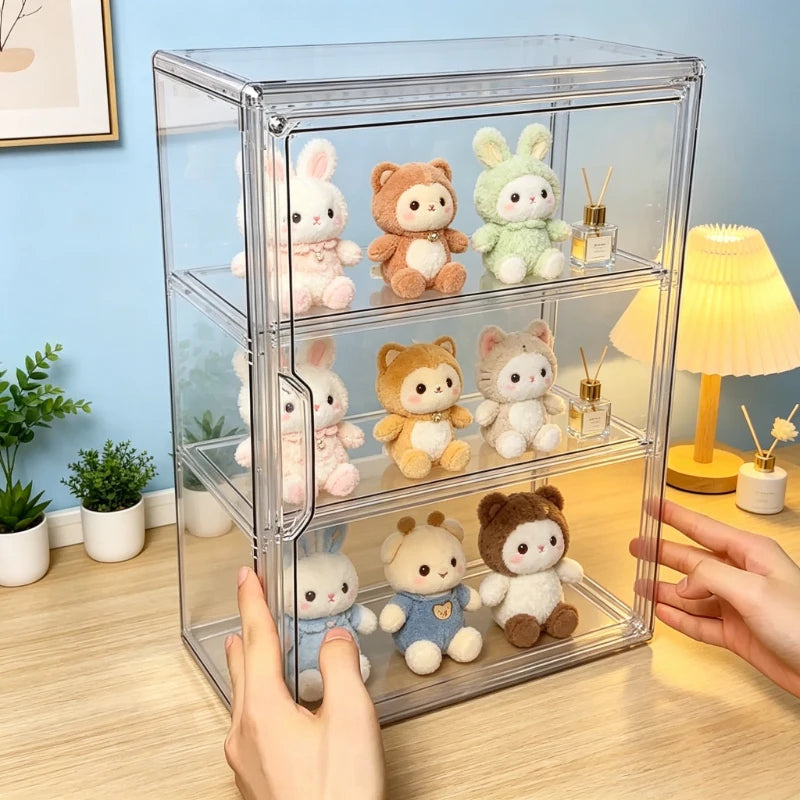 A clear acrylic three-tier display cabinet holding small plush bunny and bear toys, shown on a tabletop with hands holding the cabinet and a softly lit room in the background.