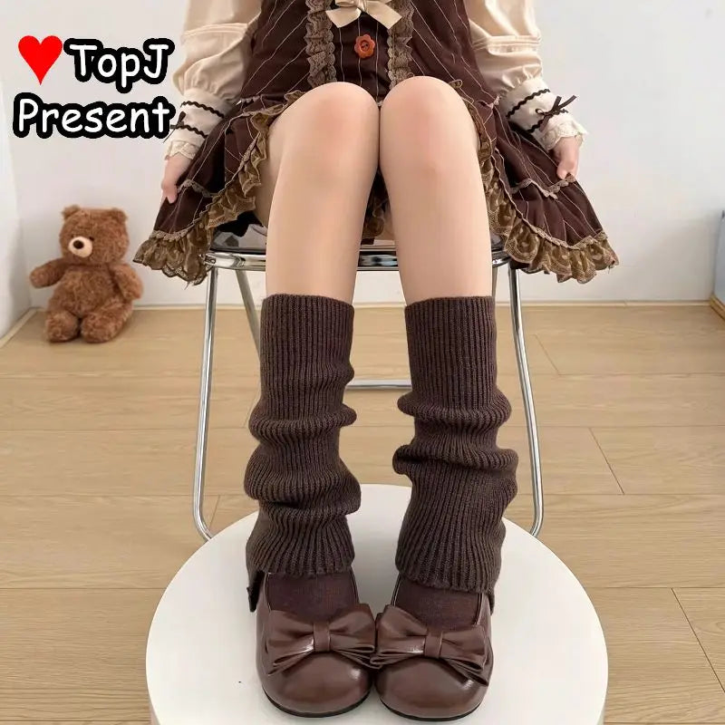A person seated on a chair wearing dark brown ribbed leg warmerss and matching brown bow flats, with a brown and beige dress visible. A teddy bear sits on the floor in the background, and a TopJ Present logo appears in the top left corner.