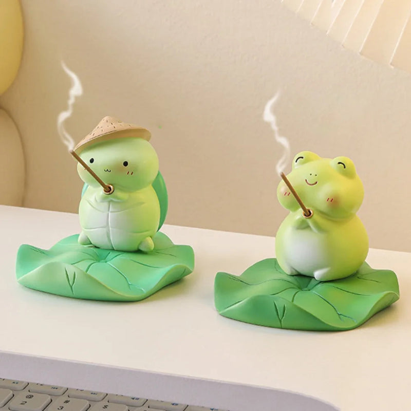 Two green frog figurines holding incense sticks on a white surface.