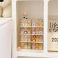 A white shelf organizer holding multiple small pastel-colored character figurines in clear plastic trays, with a plush toy partially visible on the left and a printed card on the right.