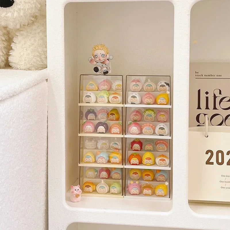 A white shelf organizer holding multiple small pastel-colored character figurines in clear plastic trays, with a plush toy partially visible on the left and a printed card on the right.