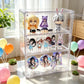 A clear acrylic three-tier display box filled with anime figurines and round character prints, styled on a bright windowsill with balloons, potted succulents, and soft natural light.