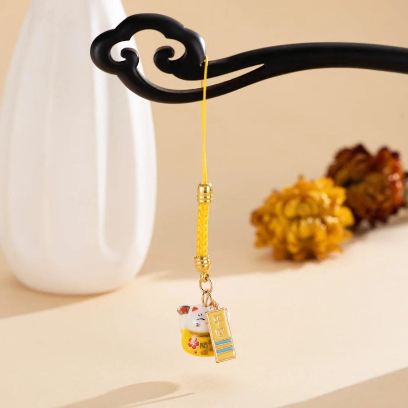 A yellow braided cord charm with gold-toned hardware and a small hanging decorative tag, displayed on a black ornamental stand against a warm beige background with soft blurred props.