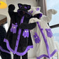 Two people wearing plush hooded pajama robes in a bedroom setting. One robe is black with purple trim and cat-ear hood details, and the other is white with purple trim and cat-ear hood details.