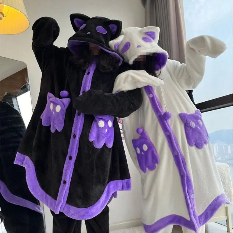 Two people wearing plush hooded pajama robes in a bedroom setting. One robe is black with purple trim and cat-ear hood details, and the other is white with purple trim and cat-ear hood details.
