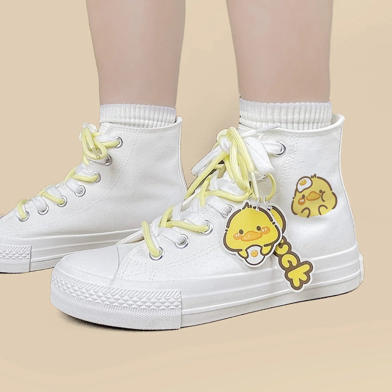 White high-top canvas sneakers with pale yellow laces and yellow cartoon duck character patches on the side, shown on a model's feet against a light beige background.