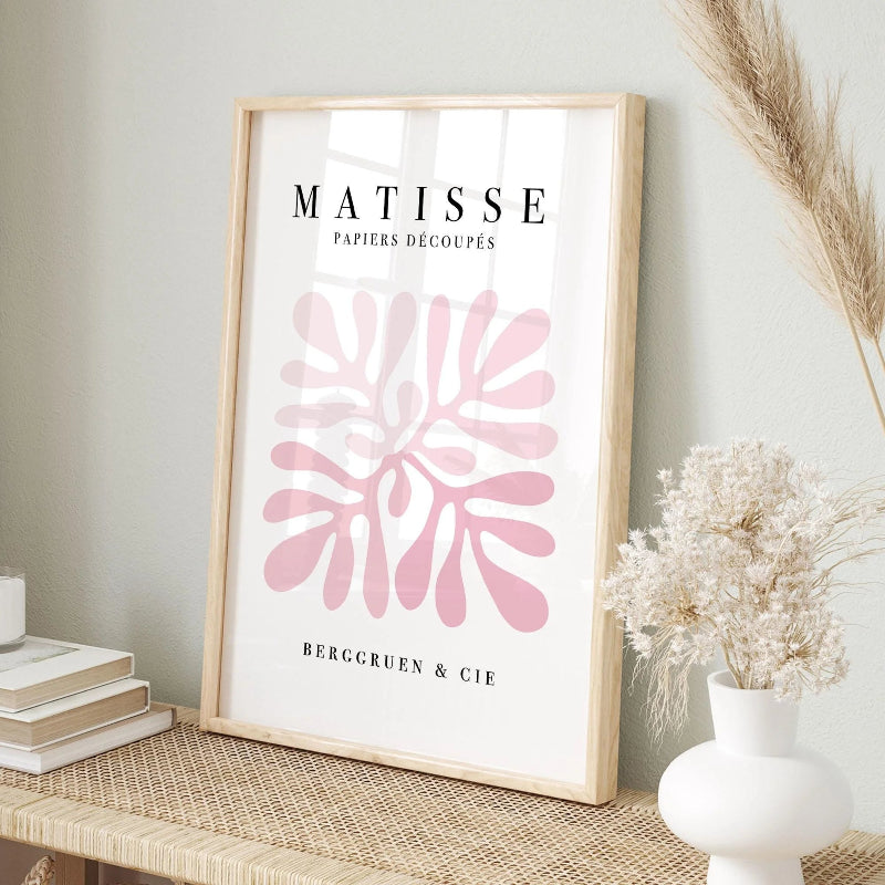 A framed art print featuring Matisse's Bergcroun & Cie decoupage paper design, styled on a woven table next to a white vase with dried grasses and a stack of books.