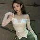Woman wearing a white lace top and green pants against a gray wall.